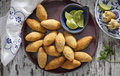 Fotografia em tons de amarelo em uma bancada de madeira com um prato redondo raso e vários bolinhos de bacalhau em cima dele. Ao lado, um potinho com fatias de limão.