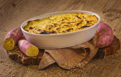 Fotografia em tons de marrom em uma bancada de madeira marrom. No centro, há um pano marrom com uma travessa branca em cima contendo o Escondidinho de Batata Doce com algumas batatas doces ao redor.