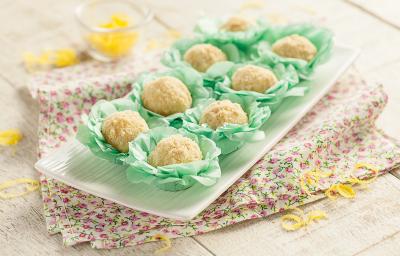 Fotografia em tons de verde em uma bancada de madeira com um recipiente branco retangular com os brigadeiros de torta de limão em forminhas verdes colocados em cima do recipiente. Ao lado, raspas de limão-siciliano espalhadas pela mesa.