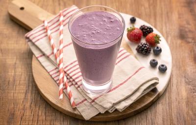 Foto em tons de roxo da receita de milkshake de uva com frutas vermelhas servida em um copo alto sobre uma tábua de madeira um pano listrado vermelho e branco com canudos de mesma cor e frutas vermelhas usadas na receita ao lado como decoração.