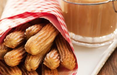 Fotografia em tons de marrom e branco de uma bancada de madeira com um prato retangular, sobre ele um paninho vermelho com churros. Ao lado um recipiente com doce de leite.