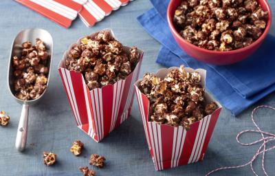 Fotografia em tons e azul e vermelho de uma bancada azul, sobre ela dois saquinhos de pipoca com pipoca de chocolate. Ao lado um pegador com pipoca, um paninho azul com um recipiente vermelho com pipoca.