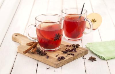 Fotografia em tons de verde em uma bancada de madeira branca, um paninho verde, uma tábua de madeira, duas xícaras de vidro com o chá de hibisco com maçã dentro deles. Ao lado, paus de canela, cravos e anis-estrelado.