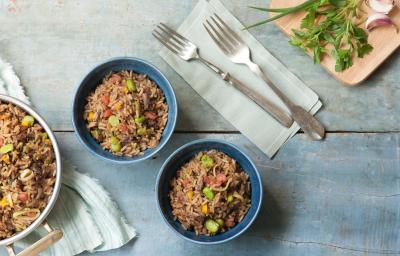 Fotografia em tons de azul em uma bancada de madeira azul com um pano branco com listras azuis, um panela com arroz e carne. Ao centro, dois potes redondos fundos com o arroz, carne e vegetais dentro dele.