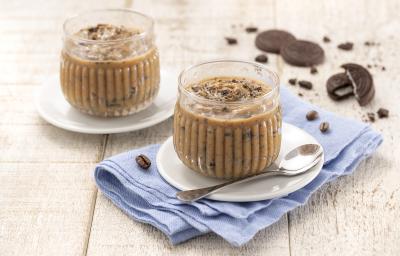 Foto da receita de Palha Italiana de Colher. Observa-se duas taças redondas e transparentes com a palha italiana de colher em um pires branco em cima de um guardanapo azul claro sobre uma superfície de madeira. Atrás, biscoitos NEGRESCO de decoração.