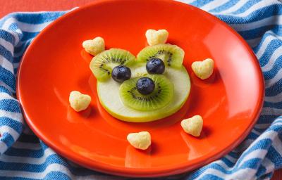 Fotografia em tons de laranja em uma bancada com paninho com listras azuis. Ao centro, um prato laranja redondo com um ursinho feito de maçã, kiwi e mirtilo e acompanhado por biscoitinhos em formato de coração. Ao lado, uma colher laranja e uma embalagem.