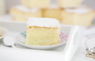 Fotografia em tons claros de um bolo de massa de pão de ló com açúcar de confeiteiro em cima, que está sobre um prato pequeno branco, com desenhos em rosa e azul. Ao fundo, mais pedaços do bolo, e ao lado do prato, colheres de café empilhadas.