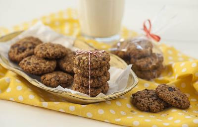 Fotografia em tons de branco e amarelo de uma bancada branca com um paninho amarelo com bolinhas brancas, sobre ele cookies. Ao fundo um copo de vidro.