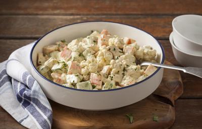 Fotografia em tons de marrom, branco e azul de uma bancada de madeira marrom com uma tábua de madeira, sobre ela um paninho branco xadrez com um recipiente redondo com a salada de legumes  e uma colher. Ao lado dois recipientes brancos.