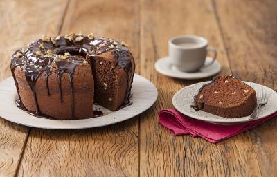 Fotografia em tons de marrom em uma mesa de madeira com um prato branco grande e o bolo de chocolate e nozes e ao lado um prato branco pequeno com uma fatia do bolo. Ao fundo, um pires e uma xícara branca.