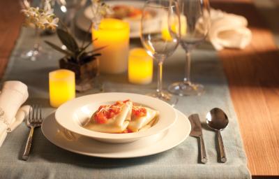 Fotografia em tons de amarelo em uma mesa de madeira e uma toalha de mesa cinza. Um sousplat branco e um prato fundo branco com a sofioli de shimeji ao molho de tomates. Velas amarelas, taças de vidro, arranjo pequeno de flor e talheres compondo a mesa.