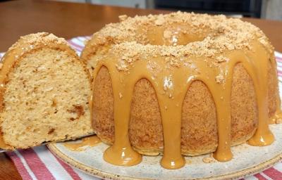 Foto em tons de caramelo da receita de bolo de paçoca com leite ninho servida em uma porção inteira com um pedaço cortado sobre uma base de cerâmica em cima de um paninho listrado rosa