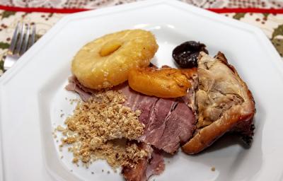 Fotografia vista de cima de um corte de carne, duas fatias de lombo, uma fatia de abacaxi assado e duas frutas assadas em um prato branco. Ao lado do prato, um garfo que está sobre uma toalha de mesa desenhada, na cor branco e verde.