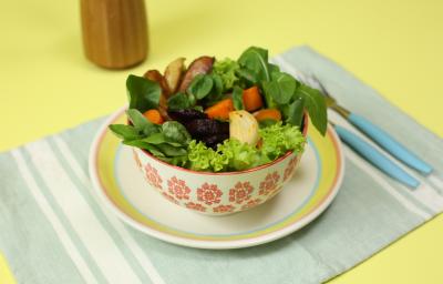 Fotografia em tons de amarelo em uma bancada de madeira amarela, um jogo americano verde claro listrado, um prato branco com listas coloridas, um potinho branco com flores e dentro uma salada de legumes.