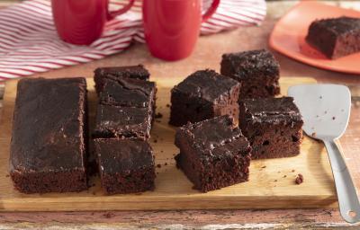 Fotografia em tons de marrom e vermelho de uma bancada de madeira, ao centro uma tábua com fatias de bolo de chocolate. Ao fundo 2 xícaras e um prato vermelho com uma fatia de bolo.