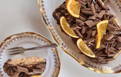 Fotografia em tons de brano com uma travessa de vidro ao centro. Em cima da travessa existe uma receita de Choco Torta coberta com raspas de chocolate ao leite e pedaços de laranja