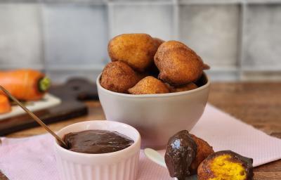 Foto da receita de bolinho de chuva servida em diversas porções em um bowl de cerâmica bege sobre um paninho rosa com um recipiente com a calda a frente. Ao fundo há uma tábua com cenouras em cima de uma mesa de madeira