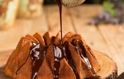 Fotografia em tons de marrom em uma bancada de madeira e ao centro uma tábua de tronco de árvore com o bolo de cenoura em cima e uma mão segurando um potinho despejando a calda de chocolate por cima do bolo.