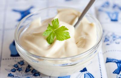 Imagem de um pequeno recipiente redondo de vidro contendo um creme branco. Uma folha de salsinha decora o centro do creme. O Recipiente está disposto sobre uma toalha em tons de branco e azul