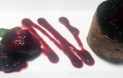 Fotografia de um flan de chocolate sobre um prato branco com uma calda de frutas vermelhas e pequenas folhas de hortelãs.