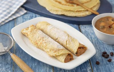 Foto da Receita de Panqueca de Doce de Leite. Observa-se um prato quadrado branco com duas panquecas em cima polvilhadas com açúcar. Do lado esquerdo, tem-se uma peneira com o açúcar e, do direito, as massas de panqueca inteiras.