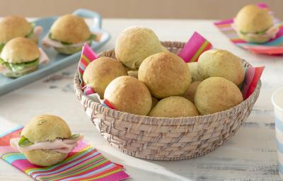 Foto da receita de Pãozinho de Talos. Observa-se uma cestinha com os mini pães dentro e, atrás, como decoração, alguns pães recheados de frios.