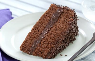 Foto de um prato branco numa mesa de madeira branca com uma fatia de bolo de chocolate dentro e um garfo do lado