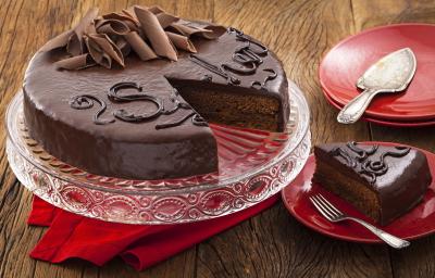 Fotografia em tons de vermelho em uma bancada de madeira com um suporte para bolo de vidro com a torta sacher de chocolate em cima cortada ao meio. Ao lado, pratinhos vermelhos e um deles com uma fatia da torta.