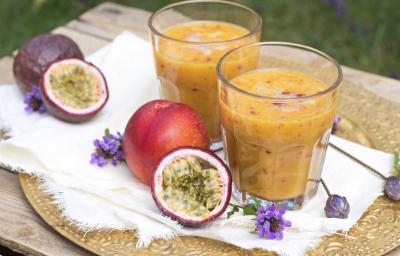 Fotografia de dois copos de vidro com uma bebida com suco de maracujá, leite e neston, apoiados sobre um pano fino branco, e por baixo, um sousplat. Ao lado dos copos, tem dois maracujás inteiros e uma metade aberta.