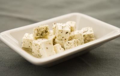 Fotografia de cubos de queijo branco fresco temperados e com azeite dentro de um recipiente pequeno, fundo e quadrado de vidro na cor branco. O aperitivo está sobre uma toalha de mesa preta.