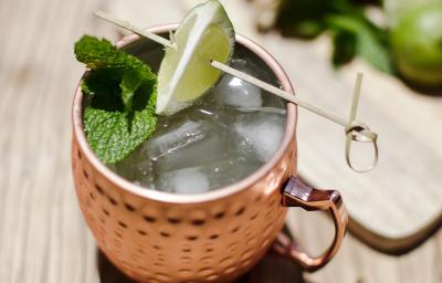 Foto da receita de Moscow Mule Blue, servida em uma caneca de estanho com gelo, sobre uma bancada de madeira decorada com algumas fatias de limão