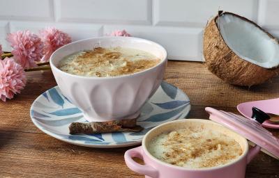 Foto da receita de cocada de panela de pressão, servida em uma tigela branca, sobre um prato decorado com pau de canela, numa bancada de madeira com um pedaço de coco seco aberto e algumas flores rosas de mentira ao fundo