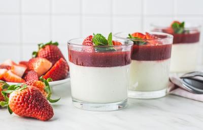 Fotografia em tons de branco em uma bancada de madeira de cor branca. Ao centro, um recipiente contendo a mousse. Ao lado, há alguns morangos espalhados e algumas colheres de inox.