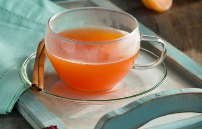 fotografia em tons de laranja, azul e cinza de uma bancada cinza vista de frente. Contém uma bandeja de madeira azul com um prato redondo transparente ao lado uma canela em pau e por cima uma xicara transparente com o chá. Ao lado um pano azul.