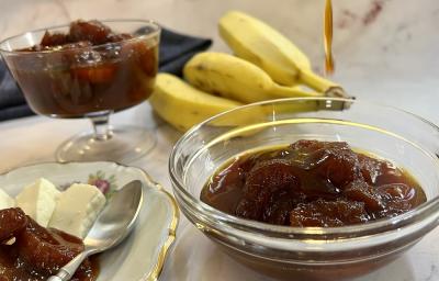 Foto da receita de compota de banana servida uma parte em um bowl de vidro e outra em uma taça baixa atrás. Ao lado, há um prato de porcelana com queijo branco e compota de banana e uma colher prateada. Ao fundo um cacho de bananas e um pano preto.