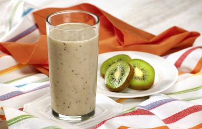 Fotografia em tons de laranja em uma bancada de madeira com uma toalha branca como listras coloridas, um pano laranja, um prato branco redondo pequeno com fatias de kiwi e um copo de vidro alto com o smoothie de iogurte com kiwi e banana.