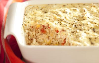 fotografia em tons de marrom, branco e vermelho de uma bancada marrom vista de frente, contém um recipiente quadrado branco com arroz e queijo por cima. Ao lado um pano vermelho com listras roxas.