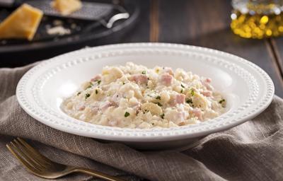 Foto em tons de cinza e branco de uma bancada de madeira escura com uma pano cinza dobrado, um garfo dourado envelhecido e uma prato redondo branco com o risoto dentro. Ao fundo, um prato preto e uma jarrinha de azeite.