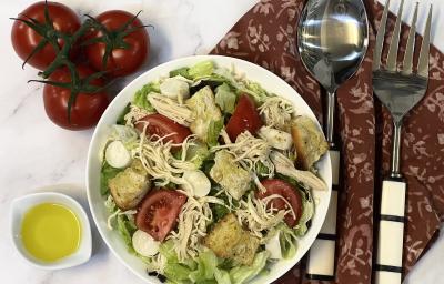 Foto vista de cima da receita de Salada de Frango servida em um recipiente grande, com um recipiente menor com Molho de Mostarda. Tudo está sobre uma bancada decorada com um tecido vermelho, um par de talheres e alguns tomates