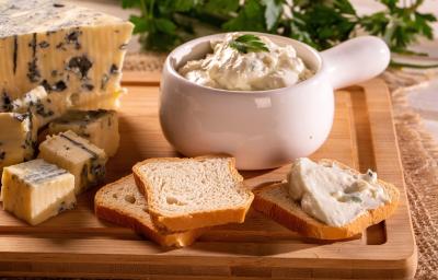 Fotografia de um recipiente pequeno de vidro branco com patê de gorgonzola de damasco, ao lado de uma peça e alguns pedaços de gorgonzola e algumas fatias de torrada, que estão sobre uma tábua de madeira.