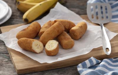 Fotografia em tons de amarelo e azul em uma bancada de madeira clara, um pano azul com listras brancas, uma tábua de madeira com várias bananas à milanesa em cima dela. Ao lado, uma concha de fritura e ao fundo um cacho de bananas.