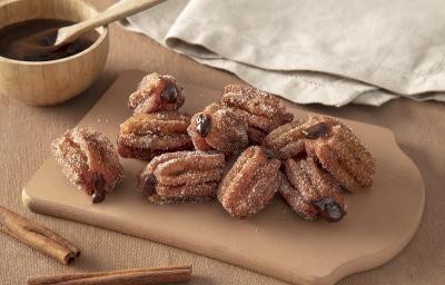 Foto em tons de bege da receita de mini churros recheados com chandelle servida em diversas porções sobre uma tábua de madeira em cima de uma mesa bege com um paninho cinza ao fundo