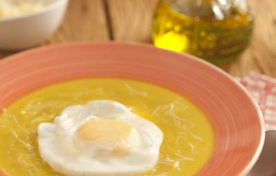 Fotografia em tons de laranja em uma bancada de madeira escura, um pano desenhado laranja, um prato laranja com a sopa portuguesa com ovo pochê dentro dele. Ao fundo, um vidrinho de azeite e um potinho com queijo ralado.