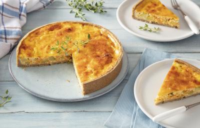 Foto em tons de branco da receita de torta de legumes com iogurte natural servida em uma porção grande sobre uma base de cerâmica com duas fatias ao lado sobre pratos menores