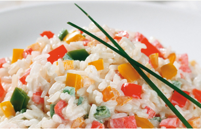 Fotografia em tons de branco e multicoloridos, porcelana branca com porção de arroz com legumes picados e decorados com duas folhas de cebolinha-verde, dois guardanapos brancos ao lado, tudo sobre bancada branca.