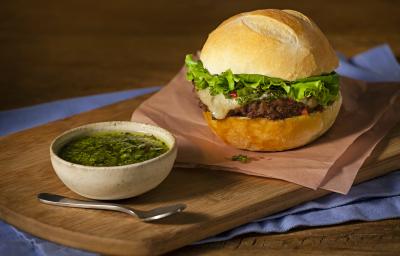 Fotografia em tons de azul em uma bancada de madeira, com uma tábua de madeira, um guardanapo marrom, um hambúrguer caseiro em cima dele. Ao lado, um potinho com molho chimichurri.