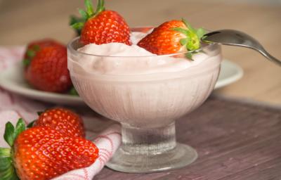 Fotografia de um creme de frutas vermelhas com iogurte, sobre o gelado, dois morangos inteiros e uma colher apoiada no recipiente de vidro em que está o creme. Ao lado, dois morangos, e ao fundo, mais morangos em cima de um prato branco pequeno.