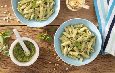 Fotografia em tons de verde em uma bancada de madeira escura, um pano branco listrado com azul, dois pratos azuis com macarrão penne com molho pesto. Ao lado, um potinho branco com o molho pesto dentro dele.