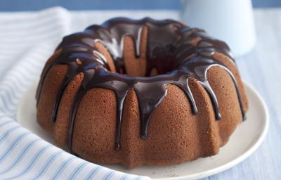 Fotografia em tons de azul em uma bancada de madeira clara com um pano branco listrado em azul, ao centro um prato branco raso com o bolo de chocolate, com cobertura de chocolate. Ao lado, um potinho com chocolate e uma jarra azul claro.