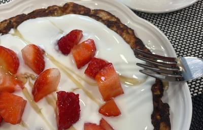 Fotografia de uma panqueca com iogurte, mel e morangos picados por cima sobre um prato raso branco de vidro, ao lado de um garfo e uma xícara branca com café.